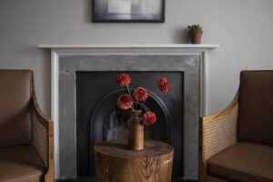 marble fireplace flanked by wooden chairs with a vase of flowers on a wooden stool